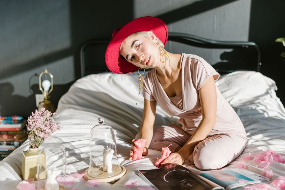 Femme en pyjama rose avec un chapeau rouge, assise sur un lit décoré de pétales de rose et feuilletant un magazine.