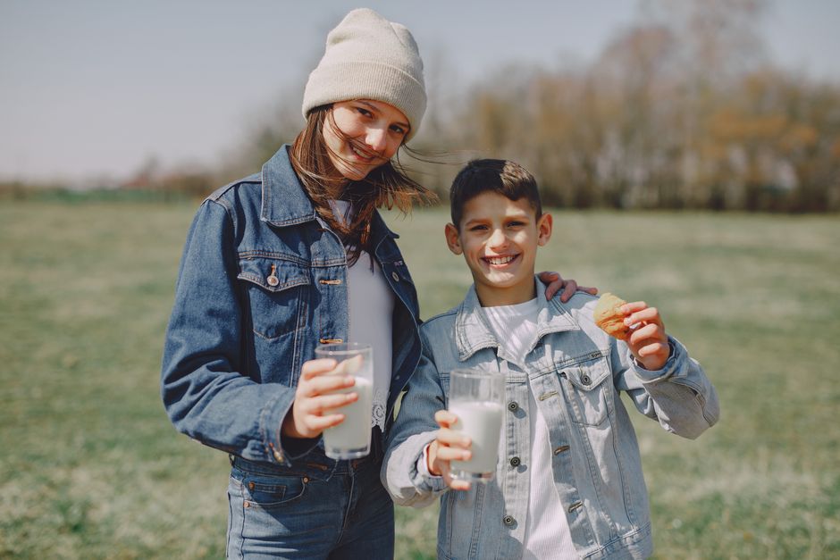 Deux enfants souriants en tenue décontractée tiennent des verres de lait et des biscuits dans un champ verdoyant.