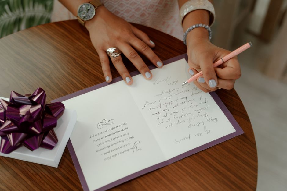 Femme écrivant un message sur une carte de vœux posée sur une table en bois, avec un cadeau décoré d'un nœud violet.