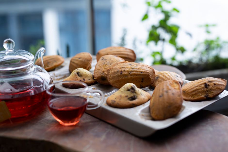 Assiette de madeleines dorées accompagnées d'une tasse de thé rouge, sur une table avec des plantes en arrière-plan.