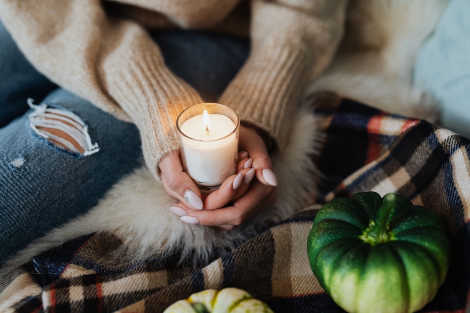 Femme tenant une bougie allumée, assise sur un plaid avec des courges décoratives à côté d'elle.