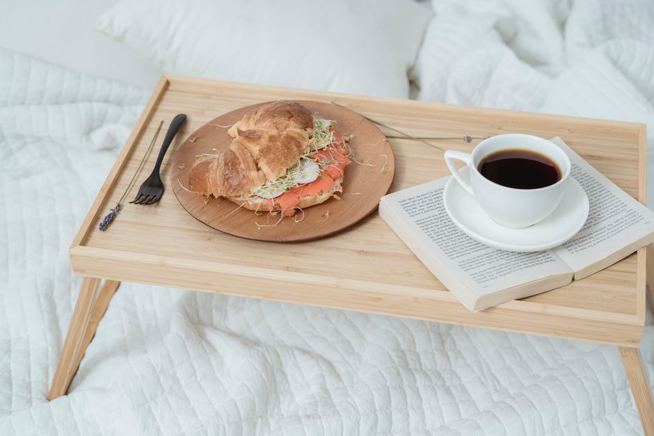 Plateau en bois avec un croissant, une tasse de café noir et un livre sur un lit avec des draps blancs.