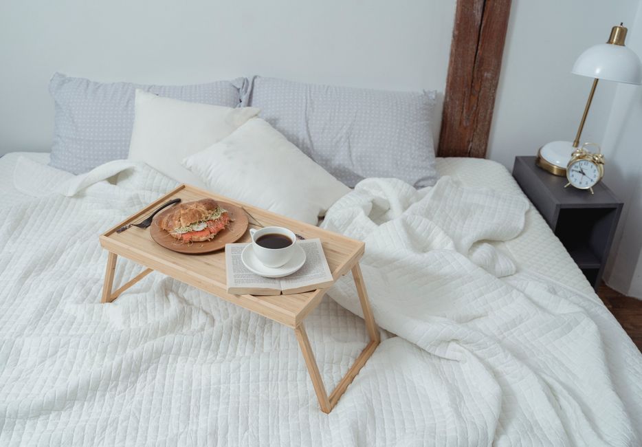 Plateau en bois avec un sandwich et une tasse de café sur un lit confortable avec des draps blancs et des coussins.