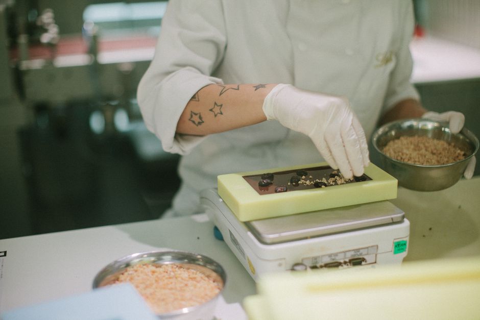 Un chocolatier prépare des bonbons en ajoutant des garnitures sur un moule, avec des ingrédients dans un bol à côté.