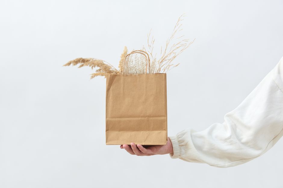 Sac en papier kraft tenu par une main, rempli de plantes séchées, sur fond blanc. Idéal pour un cadeau de dernière minute.