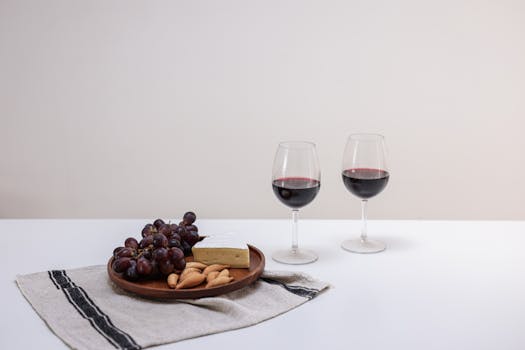 Deux verres de vin rouge, un plateau de raisins et de fromage sur une nappe en lin, évoquant la dégustation.