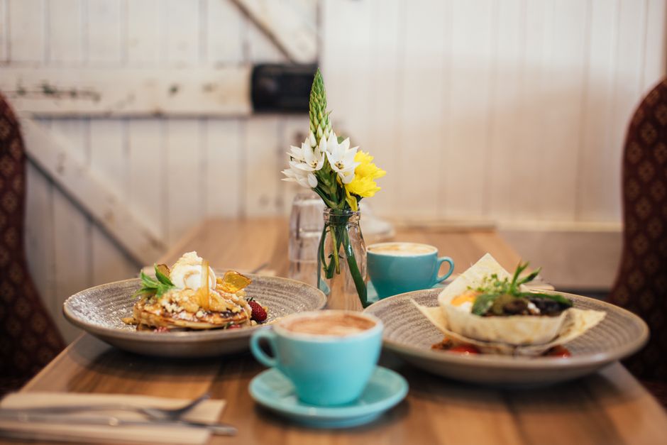 Table de brunch avec pancakes, plat salé et café, décorée d'un vase de fleurs sur un fond lumineux.