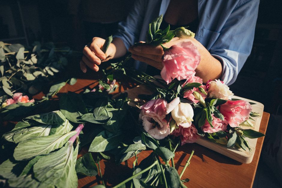 Fleuriste préparant un bouquet avec des fleurs roses et blanches sur une table en bois. Feuilles éparpillées autour.