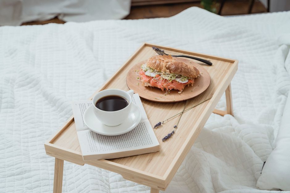 Plateau en bois sur un lit avec un croissant garni de légumes, une tasse de café et un livre. Ambiance cocooning.