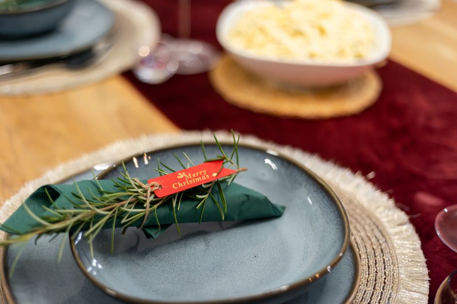 Table dressée avec assiettes bleu-gris, serviettes vertes, étiquette de Noël et plat de pâtes en arrière-plan.