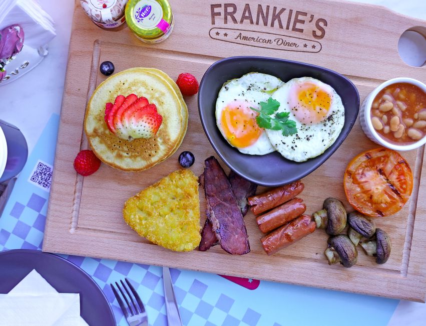 Assiette de brunch colorée avec pancakes, œufs, saucisses, haricots et fruits frais sur une planche en bois.