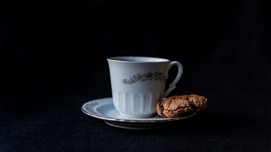 Tasse en porcelaine avec motif floral, accompagnée d'un biscuit sur une soucoupe, sur fond noir.