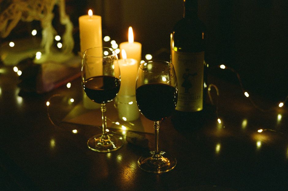 Deux verres de vin rouge posés sur une table, entourés de bougies allumées et de guirlandes lumineuses.