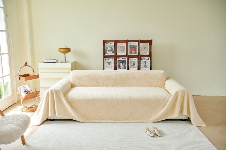 Canapé beige recouvert d'un plaid, avec des magazines en arrière-plan et des pantoufles au sol, dans une ambiance cocooning.