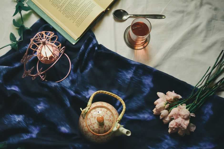 Théière en porcelaine, verre de boisson, fleurs roses et livre sur un tissu bleu, créant une ambiance romantique.