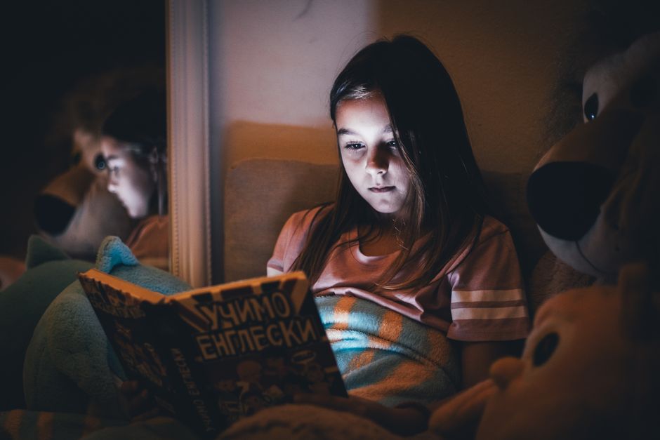 Fille lisant un livre sous une couverture, éclairée par la lumière d'un écran, dans une ambiance apaisante.