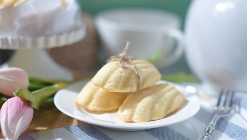 Trois madeleines dorées sur une assiette blanche, avec des fleurs et une tasse en arrière-plan.